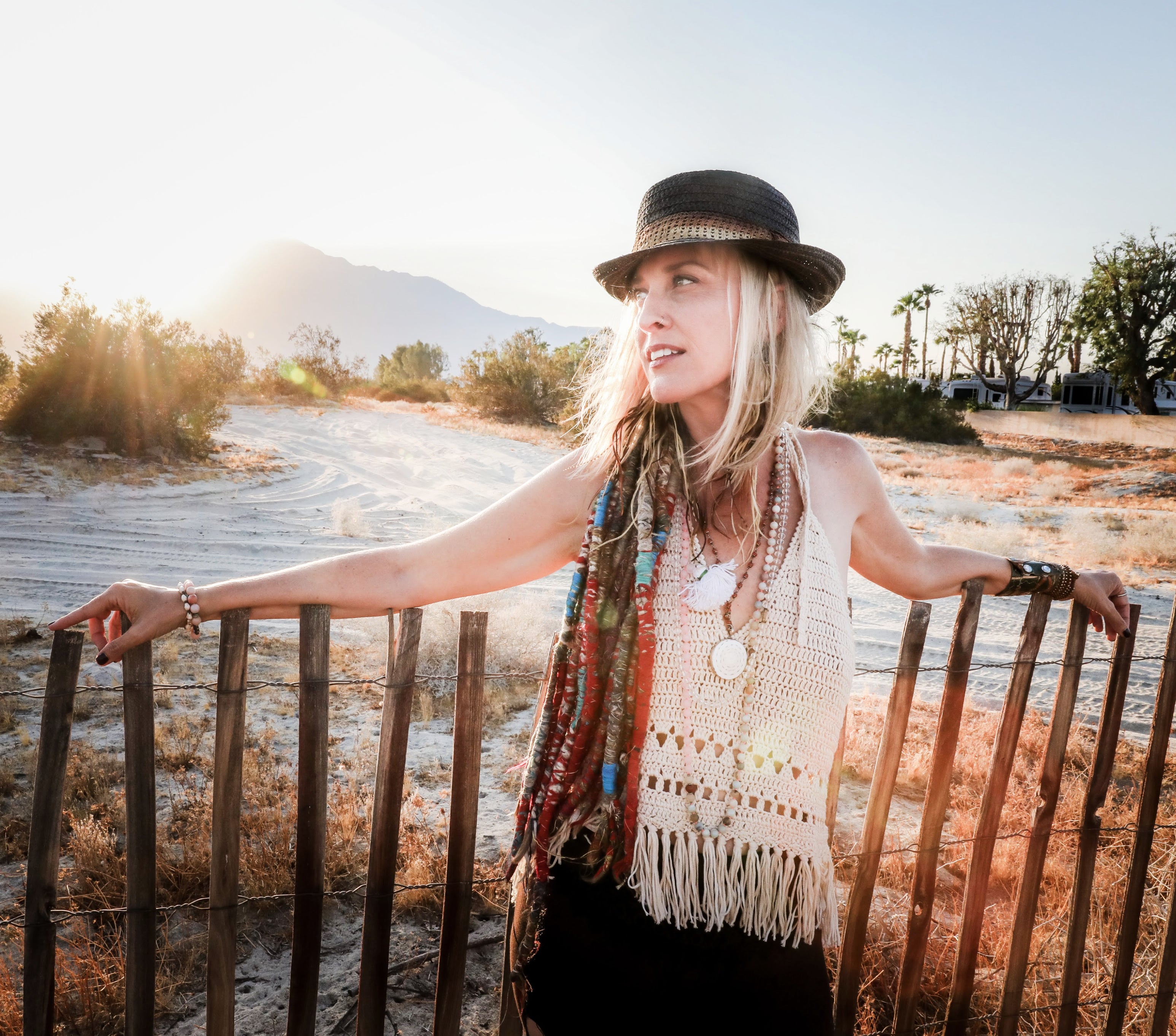 Larisa Stow stands against a rough-hewn fence in the desert with dreadlocks over her shoulder, backlit by the sun, with mountains in the distance.